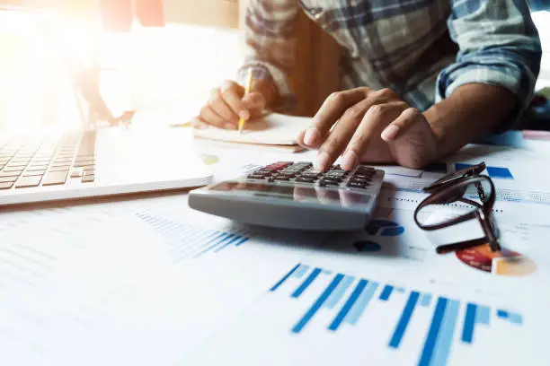 Close up of businessman or accountant hand holding pen working on calculator and laptop computer to calculate business data during make note at notepad, accountancy document at office, business financial concept