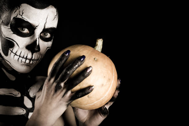 Isolated on black, closeup picture, young brunette caucasian pretty woman with scull body art with a pumpkin in hands, grey eyes, position at left, look at camera