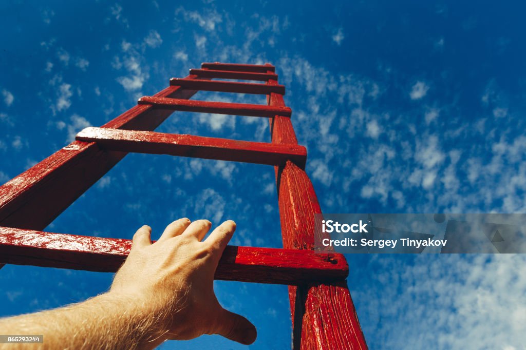 Hand Of Man Reaching For Red Ladder Leading To A Blue Sky. Development motivation Career Growth Concept A male hand holds onto the crossbar of a red wooden staircase leading to the blue sky Business Stock Photo