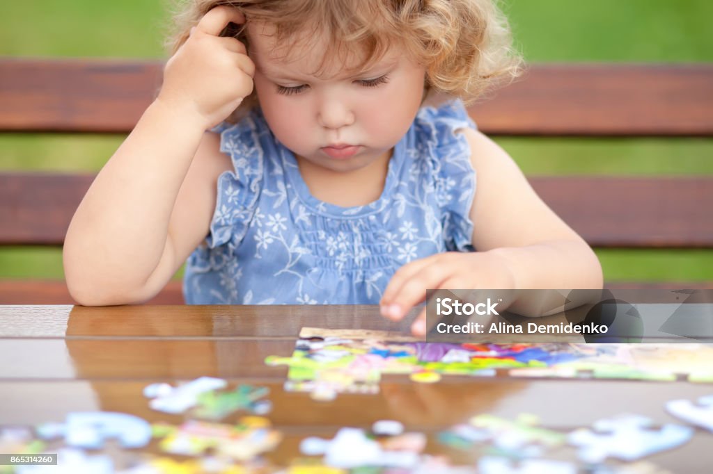 Difficult task. Tired child playing jigsaw with serious face. Blonde unhappy toddler girl, solving puzzle on a table, hard difficult task. Early education and developement. Little genius concept. Emotional. Child Stock Photo