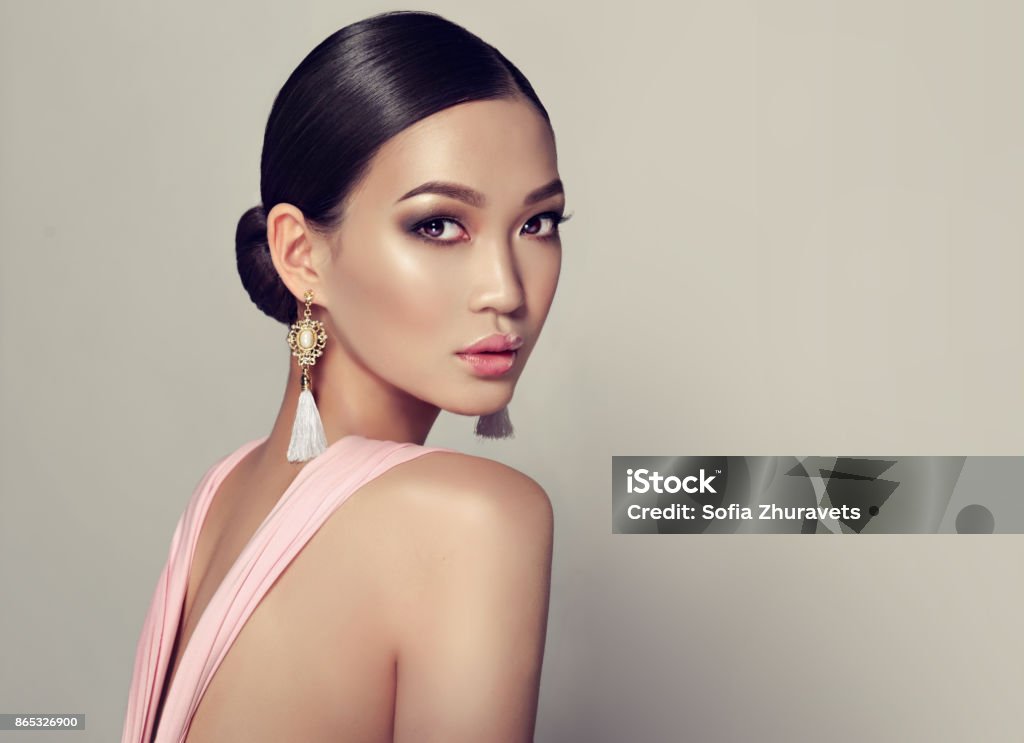 Young, gorgeous asian woman in a smoky eyes style make up, dressed in a tassel earrings. Young, gorgeous asian fashion-model put on in a smoky eyes style make up, black hair gattered in a beam. dressed in tassel earrings and  pink gown. Asian beauty. Women Stock Photo