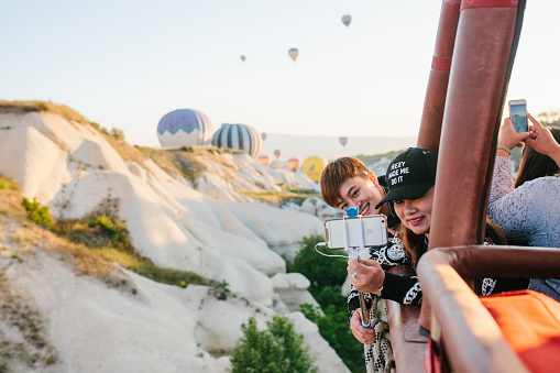 Asian tourists shoot video on iPhone during hot air balloon flight against the background of other flying balloons on sunny day