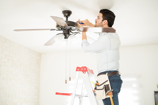 Attractive young handyman stepping on a ladder and fixing a ceiling fan