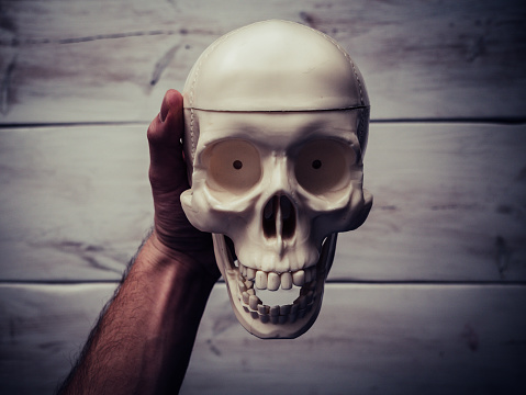 Male hand holding a human skull in the air. Wooden table in the background.