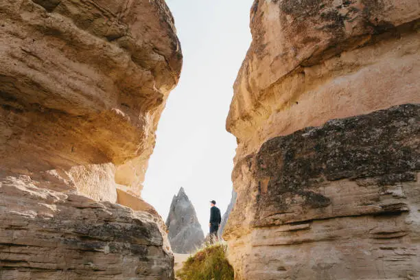 Photo of The man in dark clothes and baseball cap stands between beautiful rocks and admires the landscape in Cappadocia in Turkey