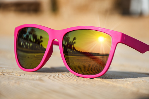 Fashionable pink sunglasses on a wooden pier with nice reflection of the beach with palm trees and sunset
