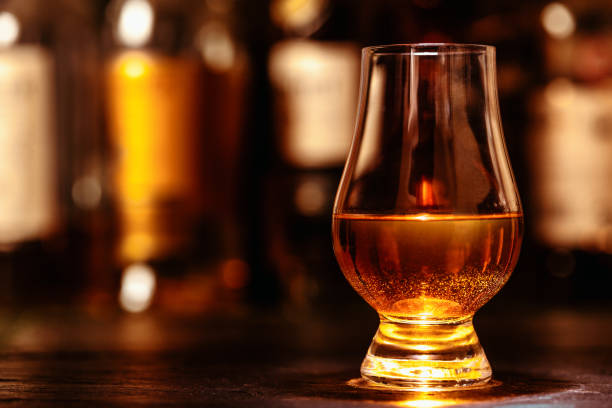 Glass of whisky on a wooden table, with a range of whisky bottles lined up in a dark background.