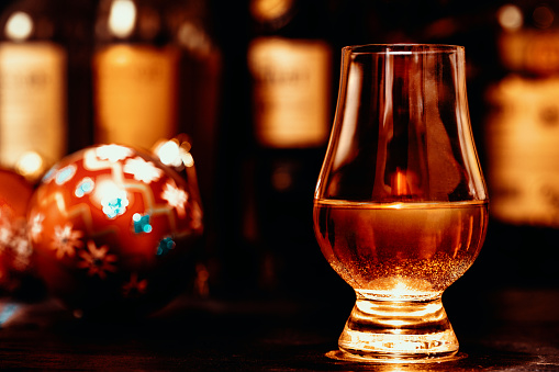 Glass of whisky on a wooden table, Christmas baubles behind, and a range of whisky bottles lined up in a dark background.