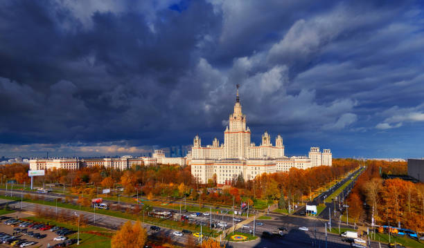 istock Gradient softened view of sunset building of Moscow university with glowing reflecting windows, red autumn trees, avenue crossing and heavy lead dramatic sky 865464562