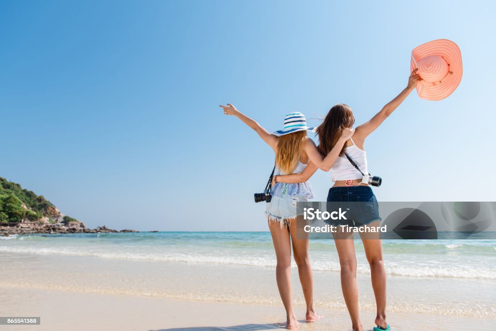 Portrait of two young asian female friends walking on the sea shore turn back at camera laughing. Multiracial young women strolling along a beach. Beach Stock Photo