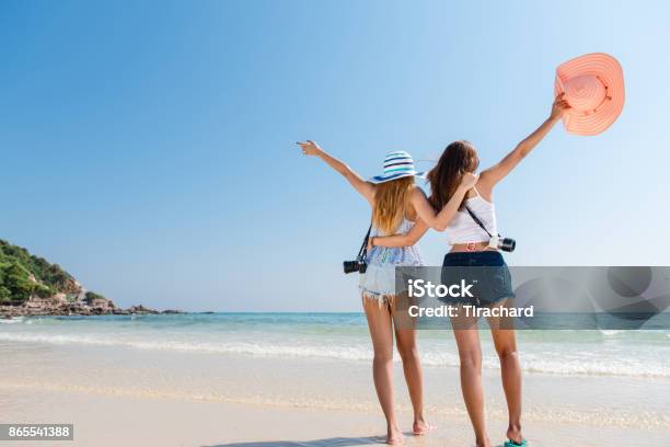 Portrait Of Two Young Asian Female Friends Walking On The Sea Shore Turn Back At Camera Laughing Multiracial Young Women Strolling Along A Beach Stock Photo - Download Image Now