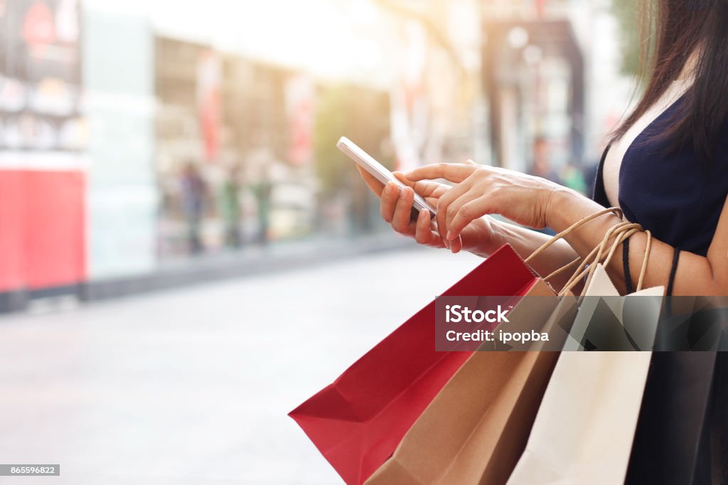 Mujer con smartphone y sosteniendo la bolsa estando en el fondo del centro comercial - Foto de stock de Venta al por menor libre de derechos