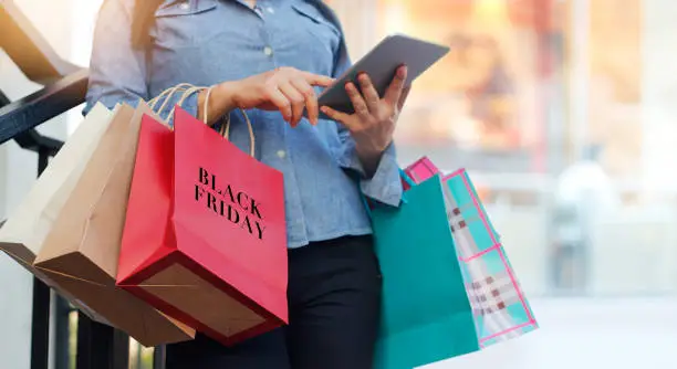 Photo of Woman using tablet and holding Black Friday shopping bag while standing on the stairs with the mall background