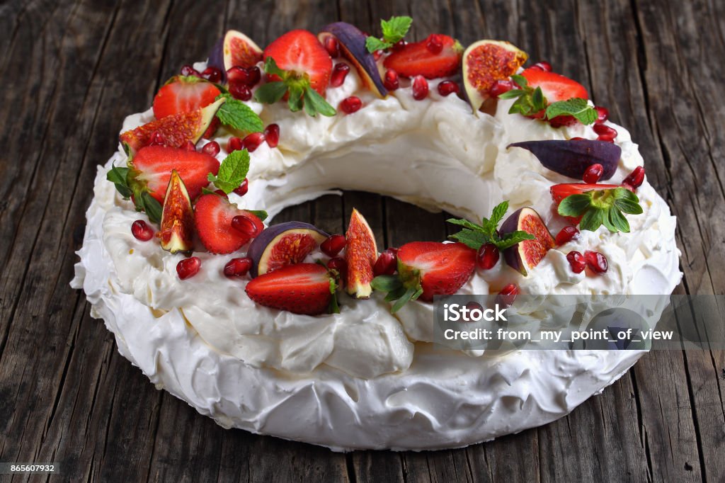 pavlova cake wreath on table, close-up pavlova cake wreath - tender and elegant sweet dessert from french meringue and whipped cream, decorated with strawberry, figs, pomegranate and mint on napkin on dark table, view from above, close-up Pavlova - Dessert Stock Photo