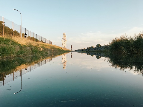 Woman Standing by Lighthouse Reflected in Big Puddle