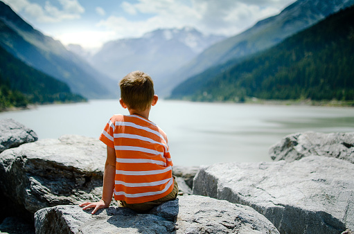 Little boy sitting with his back on the rocks looking at the mountain lake