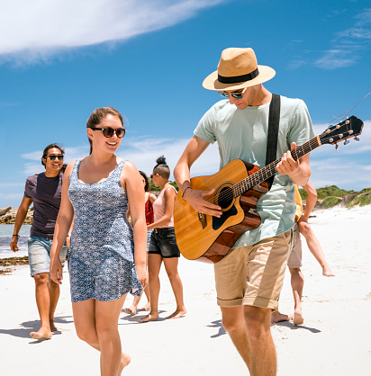 Happy woman looking at man plying guitar while walking on shore. Young males and females are enjoying summer vacation at beach. They are wearing casuals.