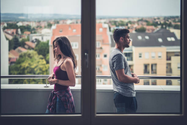 Problems in a relationship! Sad couple standing back to back on a penthouse terrace while having problems in their relationship. The view is through window. divorce couple stock pictures, royalty-free photos & images