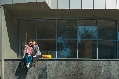 College friends take a break after classes embracing each other. Young girls with reddish hair sit on a modern building near windows on campus outdoors. Friendship and studentship.