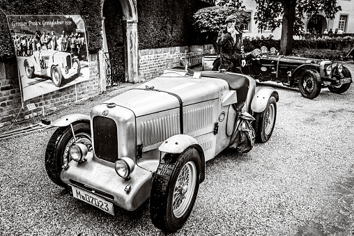 MG Molders Magnette vintage British racing car with an Alvis 12/70 sports car in the background. A man in retro style racing driver's clothing is smoking a cigarette in between the cars. The car is on display during the 2017 Classic Days event at Schloss Dyck
