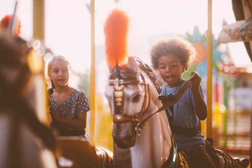 Multi-ethnic mixed family siblings having fun riding horses on funfair carousel ride in summer