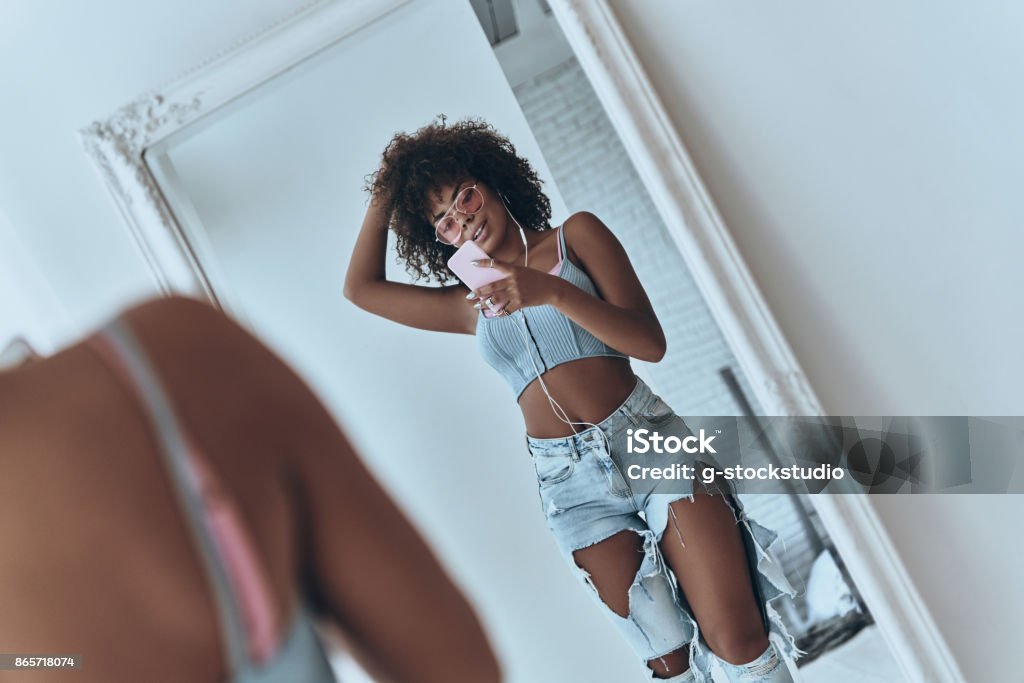 Girl with a great style. Reflection of beautiful young African woman listening music and keeping hand in hair while taking selfie in the mirror at home Mirror - Object Stock Photo