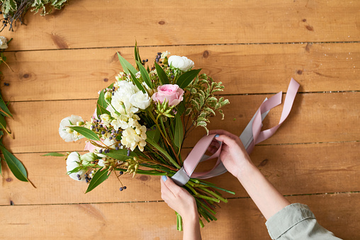 Directly above view of unrecognizable florist making floral arrangement and finishing bouquet wrapping it in ribbons on wooden table