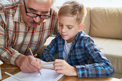Concentrated intelligent grandfather doing geometry task together with grandson and explaining sketch in workbook, home education concept