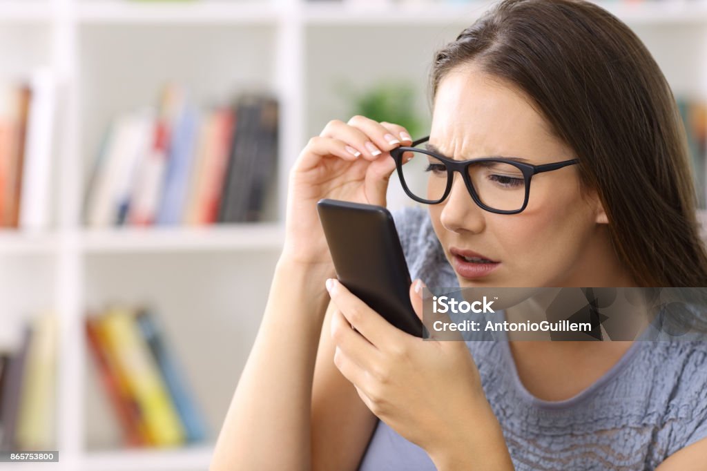 Girl with eyesight problems trying to read phone text Girl wearing eyeglasses with eyesight problems trying to read phone text at home Problems Stock Photo