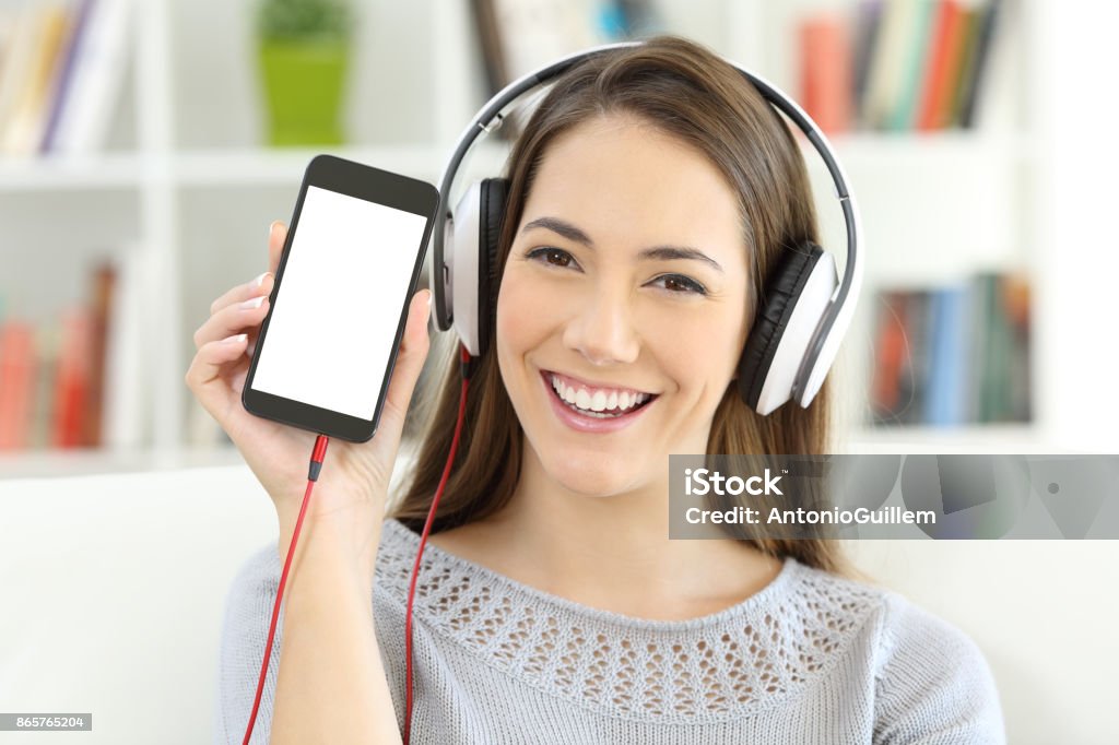 Girl listening to music showing   blank smart phone screen Front view portrait of a happy girl listening to music and showing a blank smart phone screen at home Device Screen Stock Photo