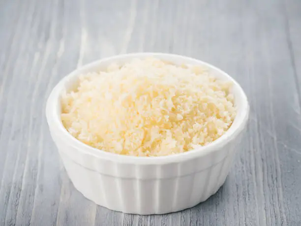 White ceramic bowl with freshly grated parmesan cheese on gray wooden background. Shredded parmesan on grey wooden table.