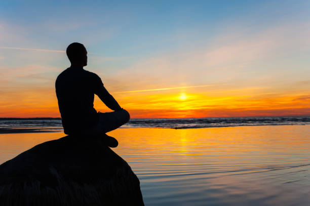 istock Man silhouette sitting on stone at the seaside contemplating sunset 865804986