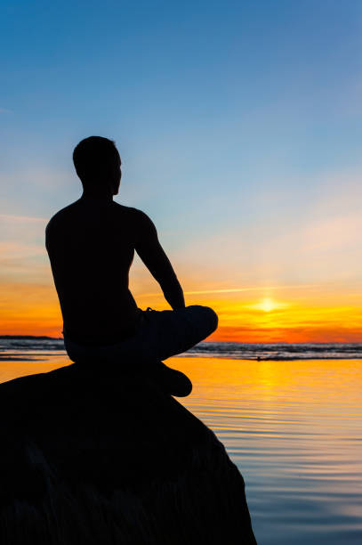 istock Man silhouette sitting on stone at the seaside contemplating sunset 865805098