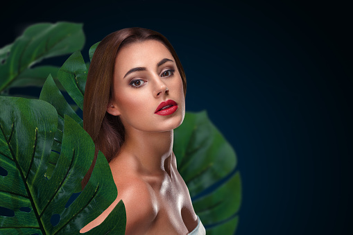 Beauty portrait of beautiful young woman with perfect skin within tropical foliage over dark background