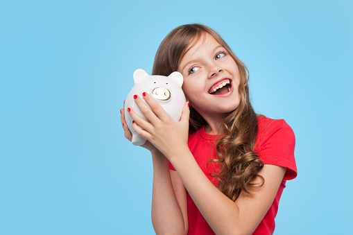 Little adorable girl holding moneybox near ear looking happy on blue background.