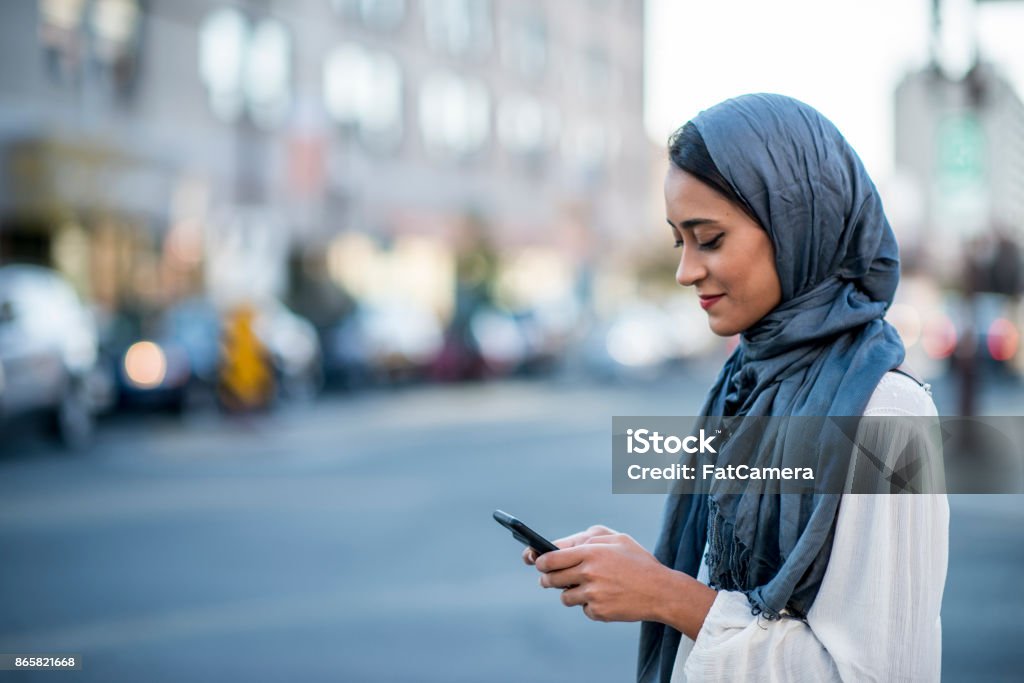 Using Technology A Muslim woman is outdoors on a sunny day. She is wearing casual clothing and a head scarf. She is standing near a road and sending a message with her smartphone. Women Stock Photo