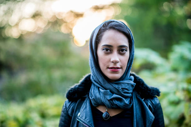 A Muslim woman is outdoors on a sunny day. She is wearing casual clothing and a head scarf. She is smiling at the camera while in a public park.