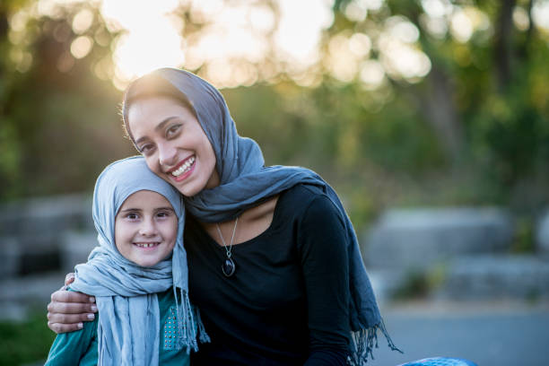 Happy Family A Muslim woman and her daughter are outdoors on a sunny day. They are wearing casual clothes and head scarves. They are sitting on a boulder in a public park, and smiling at the camera. Canada Visa stock pictures, royalty-free photos & images