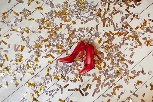 Pair of red high-heeled stylish female shoes on white wooden floor covered by confetti