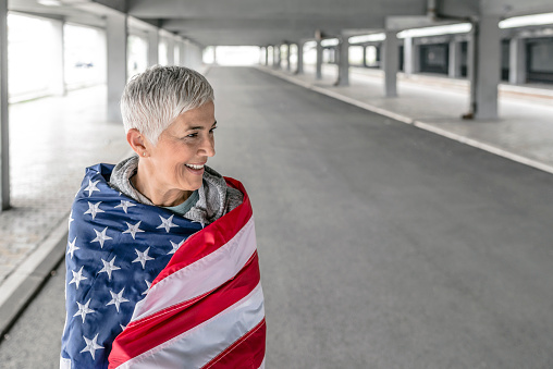 Gray hair patriotic mature woman posing in an outdoor environment,  looking forward to 4th July. Beautiful senior female wrapped in American flag. American woman holding the USA flag over a parking lot background. Happy short hair female wearing casual clothes standing with american flag.