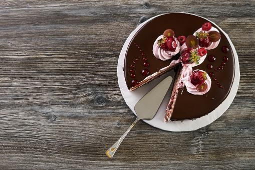 Top view of berry fruit cake with mixed fruits raspberry strawberry grapes in chocolate glaze and whipped cream and cake spatula on white cake board on wooden table background