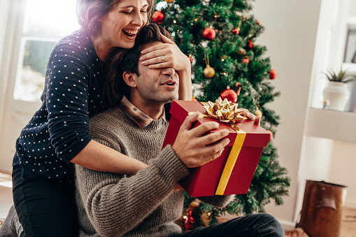 Couple spending happy moments while celebrating Christmas at home. Smiling young woman covering eyes of her partner while handing over a Christmas gift.