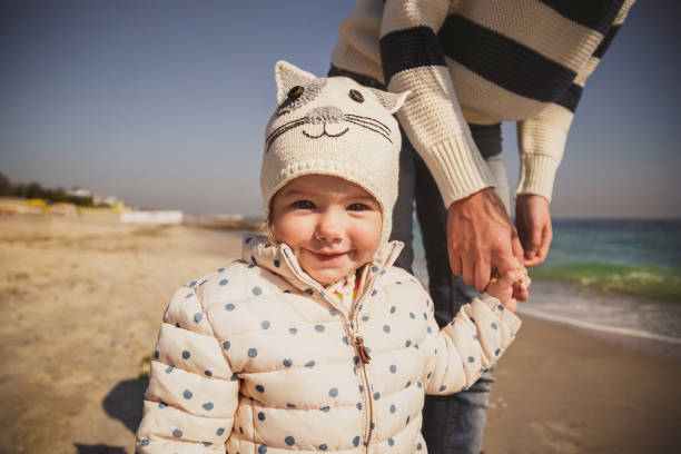 zbliżenie portret małego uśmiechniętego dziecka trzymającego rękę matki na zewnątrz oceanu, patrzącego na kamerę - winter child coat baby girls zdjęcia i obrazy z banku zdjęć