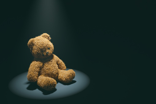 Teddy bear with torn eye illuminated by spotlight sits in dark room.