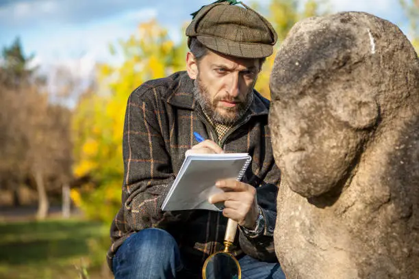 Scientists with a beard in a plaid jacket sits and describes the stone monument to the Scythian period. The left hand holds big magnifier