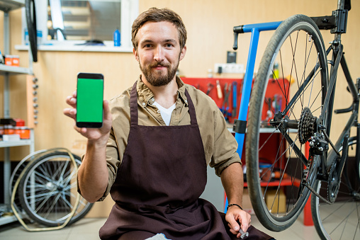 Portrait of smiling bearded mechanic sitting on haunches and showing smartphone with blank screen to camera, interior of modern bicycle repair shop on background