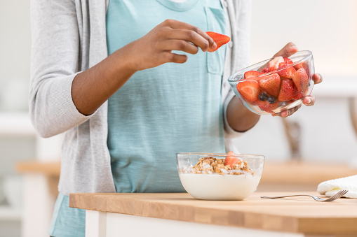 An unrecognizable woman stands at her kitchen counter and holds a bowl of strawberries.  She places one in another bowl filled with granola and yogurt, preparing a parfait for herself.