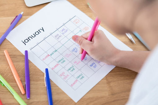 An unrecognizable  woman leans over and table and adds multiple weekly goals to a calendar for the month of January with a light blue pen.  The calendar is sitting on a wooden surface along with a note pad and other colored pens.