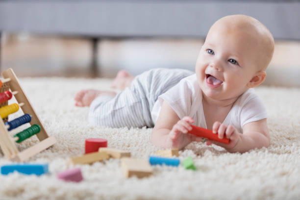 Cute baby sings with open mouth while playing with wooden blocks A cute baby lays on her tummy on a rug in a living room and sings with an open mouthed smile.  She is holding one of several wooden blocks.  There is an abacus in the foreground. toy block photos stock pictures, royalty-free photos & images