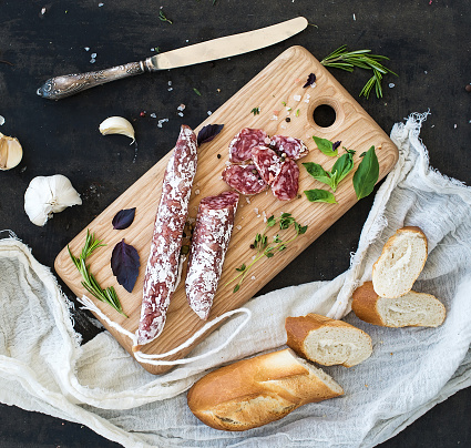 Meat gourmet snack. Salami, garlic, baguette and herbs on rustic wooden board over dark grunge backdrop, top view
